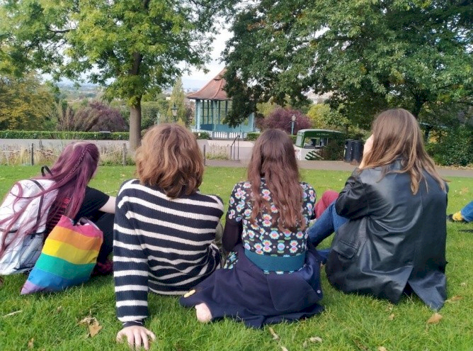 Four young people are sitting on the grass, with their backs to the camera. In the background, there are trees.
