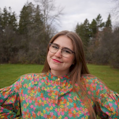 Headshot of Julia, the London Children and Youth Development Worker. She has long brown hair, wearing glasses, smiling broadly and wearing a colourful floral blouse. She is standing outside surrounded by grass and trees.